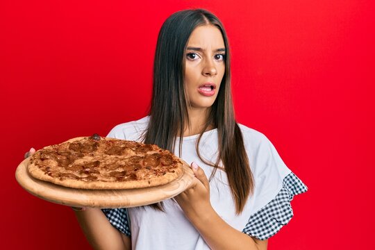Young hispanic woman holding italian pizza clueless and confused expression. doubt concept.