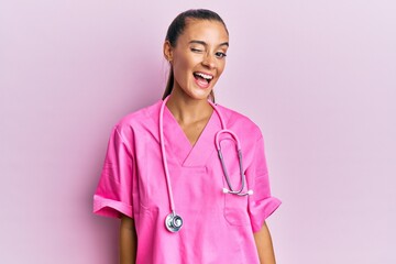 Young hispanic woman wearing doctor uniform and stethoscope winking looking at the camera with sexy expression, cheerful and happy face.