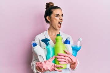 Beautiful brunette young woman wearing cleaner apron holding cleaning products angry and mad...