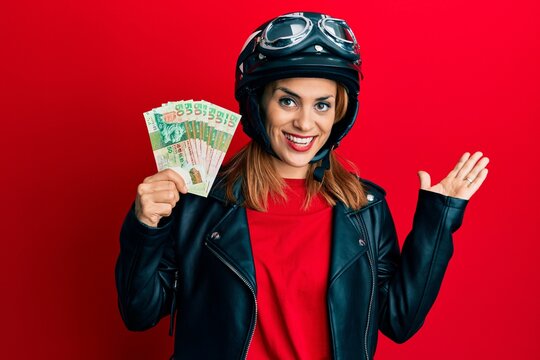 Hispanic young woman wearing motorcycle helmet holding hong kong dollars celebrating achievement with happy smile and winner expression with raised hand