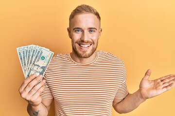 Young caucasian man holding dollars celebrating achievement with happy smile and winner expression with raised hand