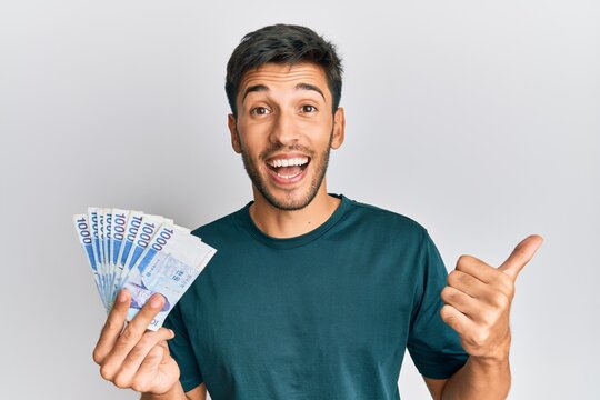 Young handsome man holding 1000 south korean won banknotes pointing thumb up to the side smiling happy with open mouth