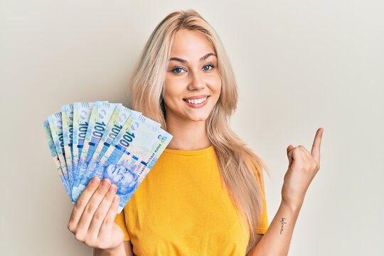 Beautiful caucasian blonde girl holding south african 100 rand banknotes smiling happy pointing with hand and finger to the side