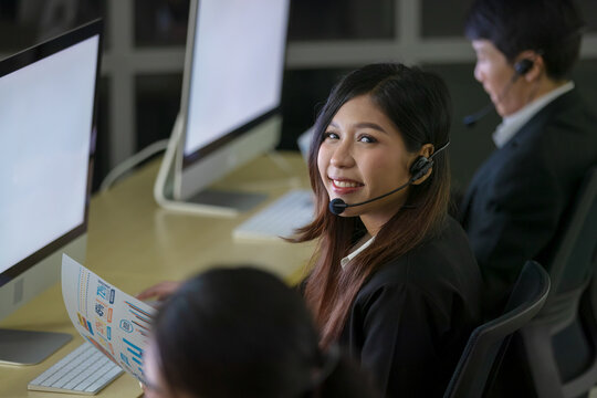 Asian smile woman wearing headphone headset and working with happy face and sevice mind in call center in front of computer screen in office at night. Overtime, late work and 24 hours service concept