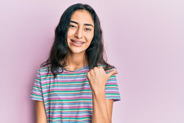 Hispanic teenager girl with dental braces wearing casual clothes smiling with happy face looking and pointing to the side with thumb up.