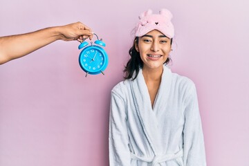 Hispanic teenager girl with dental braces wearing sleep mask and robe with alarm clock close to her with a happy and cool smile on face. lucky person.
