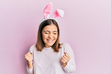 Young beautiful woman wearing cute easter bunny ears excited for success with arms raised and eyes...