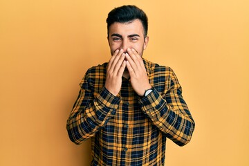 Young hispanic man wearing casual clothes laughing and embarrassed giggle covering mouth with...