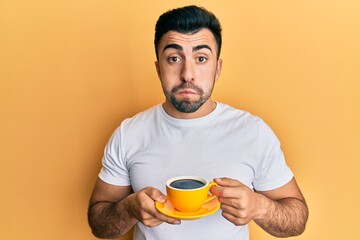 Young hispanic man drinking a cup of coffee puffing cheeks with funny face. mouth inflated with air, catching air.