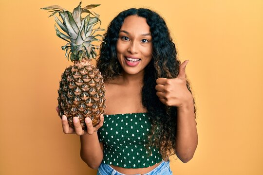 Young latin woman holding pineapple smiling happy and positive, thumb up doing excellent and approval sign