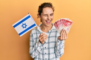Young brunette woman holding israel flag and shekels banknotes winking looking at the camera with...