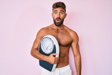Young hispanic man standing shirtless holding weighing machine scared and amazed with open mouth...