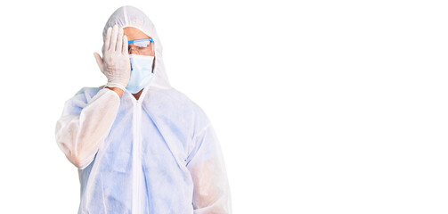 Young hispanic man wearing doctor protection coronavirus uniform and medical mask covering one eye with hand, confident smile on face and surprise emotion.