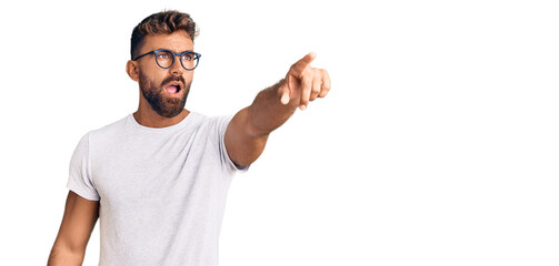 Young hispanic man wearing casual clothes and glasses pointing with finger surprised ahead, open mouth amazed expression, something on the front