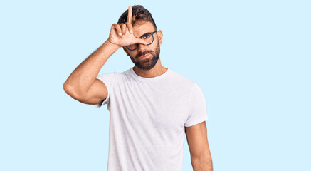 Young hispanic man wearing casual clothes and glasses making fun of people with fingers on forehead doing loser gesture mocking and insulting.