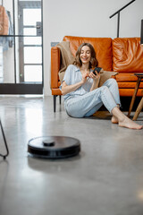 Black robotic vacuum cleaner cleaning the floor while woman sitting near sofa and using phone....