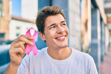 Young caucasian man smiling happy holding pink breast cancer ribbon at the city.