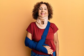 Beautiful middle age mature woman wearing cervical collar and arm on sling looking positive and...