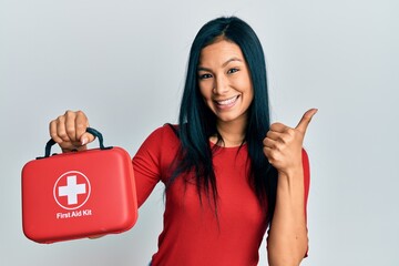 Beautiful hispanic woman holding first aid kit smiling happy and positive, thumb up doing excellent...