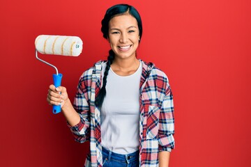 Beautiful hispanic woman holding roller painter looking positive and happy standing and smiling...