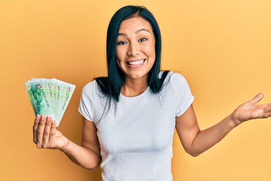 Beautiful hispanic woman holding 5 singapore dollars banknotes celebrating achievement with happy smile and winner expression with raised hand