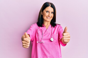 Middle age brunette woman wearing doctor uniform and stethoscope approving doing positive gesture...