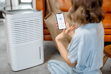 Woman sitting near air purifier and moisturizer appliance near sofa monitoring air quality in...