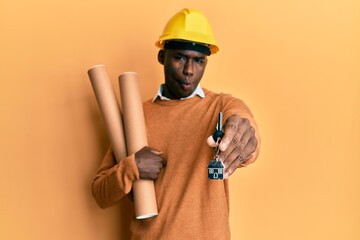 Young african american man wearing safety helmet holding blueprints and new home key making fish face with mouth and squinting eyes, crazy and comical.