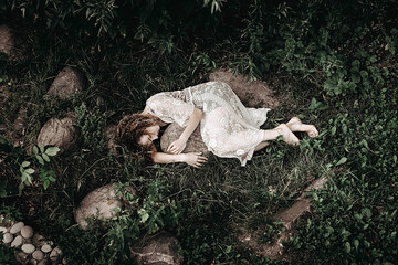 Young lonely woman laying on green grass in white mantle and hugging a huge stone. A mysterious...