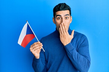 Handsome hispanic man holding czech republic flag covering mouth with hand, shocked and afraid for mistake. surprised expression