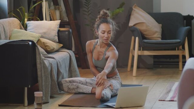 Medium tilt down shot of young woman in sportswear watching online workout and stretching on mat at home