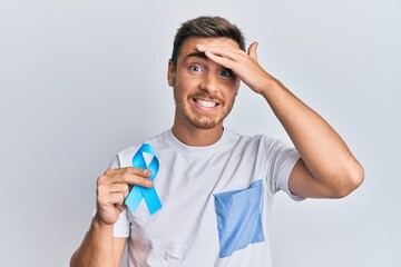 Handsome caucasian man holding blue ribbon stressed and frustrated with hand on head, surprised and...