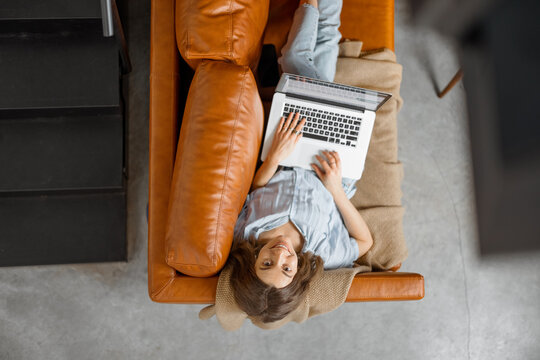 Young woman working with laptop while lying on foxy leather sofa and looking at camera. Digital nomad and work from home concept. Top view