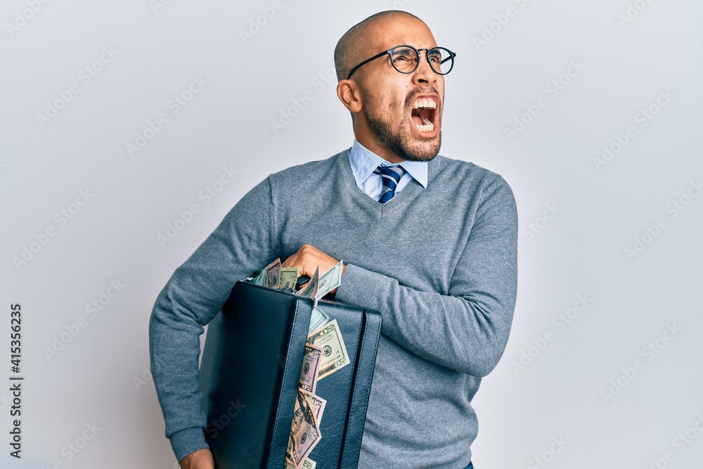 Wall mural Hispanic adult man holding briefcase full of dollars angry and mad screaming frustrated and furious, shouting with anger looking up.