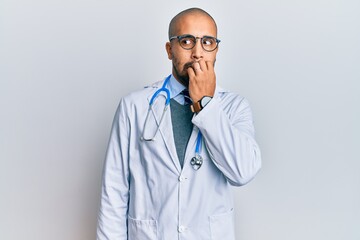 Hispanic adult man wearing doctor uniform and stethoscope looking stressed and nervous with hands on mouth biting nails. anxiety problem.