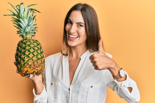 Young beautiful woman holding pineapple smiling happy and positive, thumb up doing excellent and approval sign