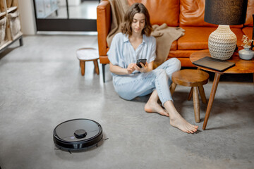 Black robotic vacuum cleaner cleaning the floor while woman sitting near sofa and using phone....