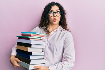 Young brunette woman with curly hair holding a pile of books making fish face with mouth and squinting eyes, crazy and comical.