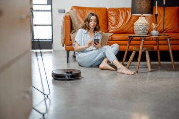 Black robotic vacuum cleaner cleaning the floor while woman sitting near sofa and using phone....