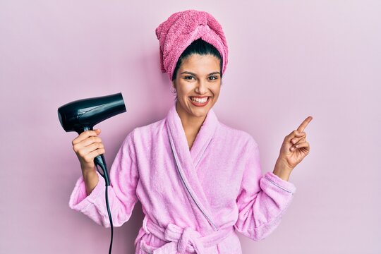 Young hispanic woman wearing shower bathrobe using dryer smiling happy pointing with hand and finger to the side