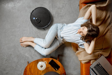 Black robotic vacuum cleaner cleaning the floor while woman sitting near sofa and relax. Top view....