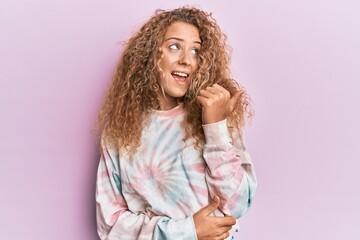 Beautiful caucasian teenager girl wearing casual tie dye sweatshirt smiling with happy face looking and pointing to the side with thumb up.