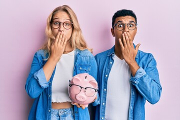 Young interracial couple holding piggy bank with glasses covering mouth with hand, shocked and afraid for mistake. surprised expression