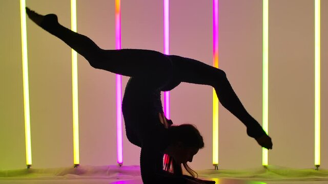 Young woman doing gymnastic exercises in the studio against the background of multicolored neon tubes. Close up of a girl's legs moving in the air, who performs a handstand. Silhouette. Slow motion.