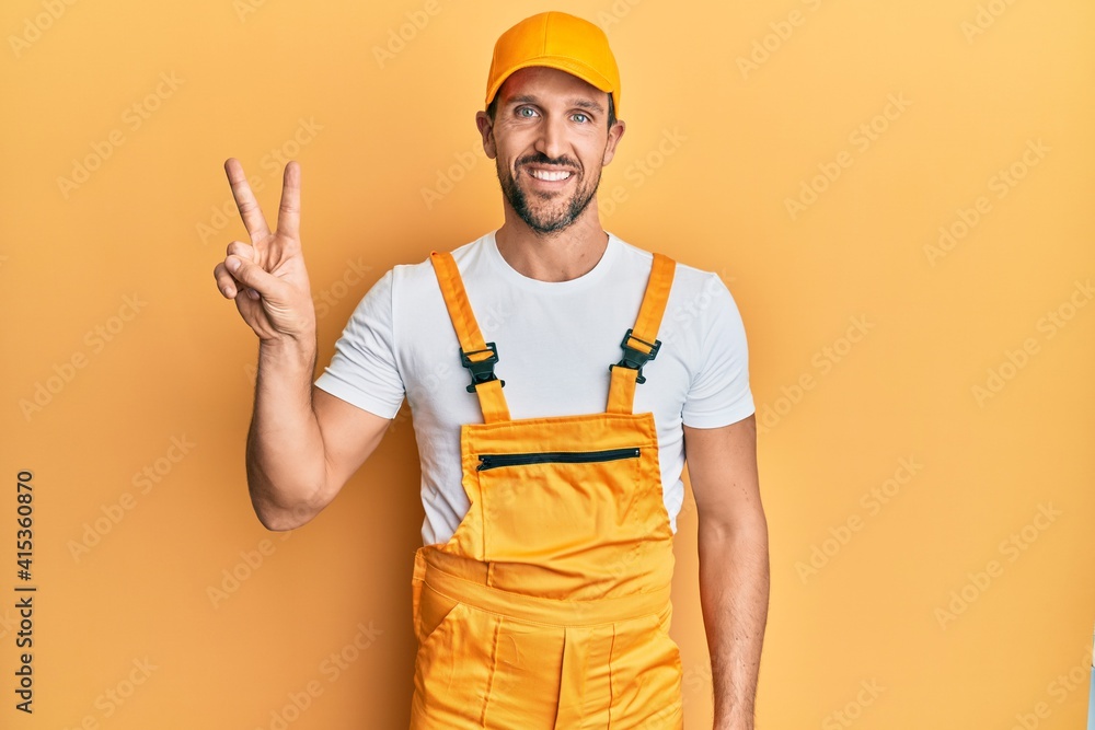 Wall mural Young handsome man wearing handyman uniform over yellow background smiling looking to the camera showing fingers doing victory sign. number two.