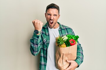 Handsome man with beard holding paper bag with groceries angry and mad raising fist frustrated and...