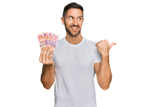 Handsome man with beard holding south african 50 rand banknotes pointing thumb up to the side smiling happy with open mouth