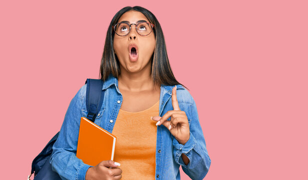 Young latin girl wearing student backpack and holding books amazed and surprised looking up and pointing with fingers and raised arms.