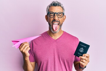 Middle age grey-haired man holding paper plane and passport sticking tongue out happy with funny...