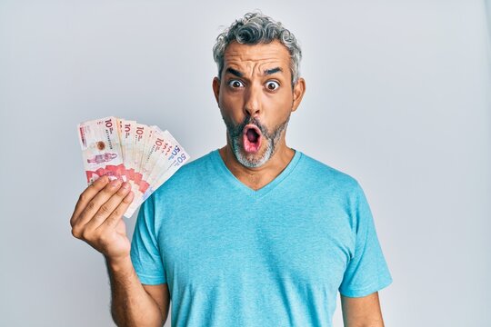 Middle age grey-haired man holding colombian pesos scared and amazed with open mouth for surprise, disbelief face